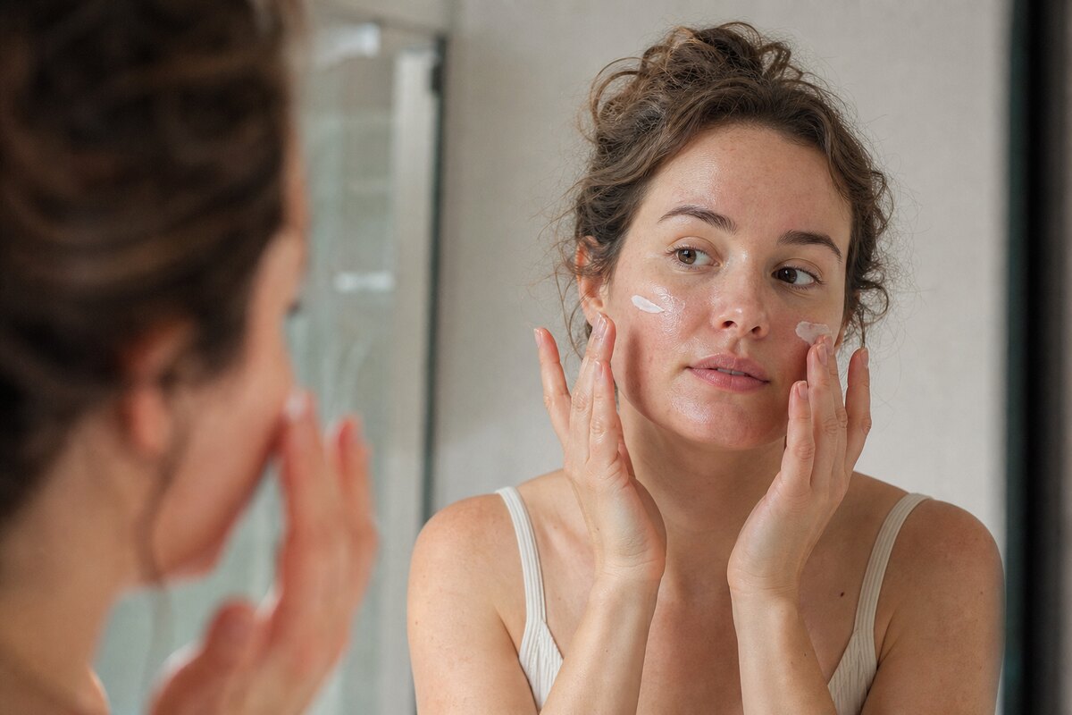 American woman applying facial sunscreen to her cheeks in front of a bathroom mirror